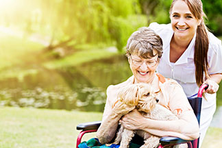 Elderly woman in wheelchair with small dog on lap, assisted by caregiver near a pond.