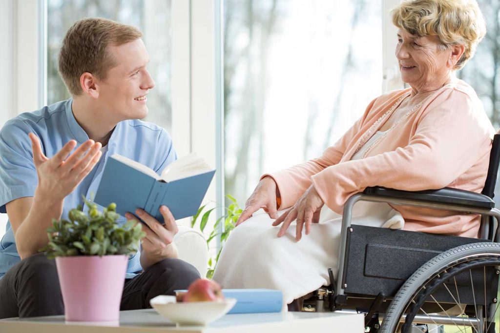 Caregiver reading to a senior woman in a wheelchair beside a plant on a table.