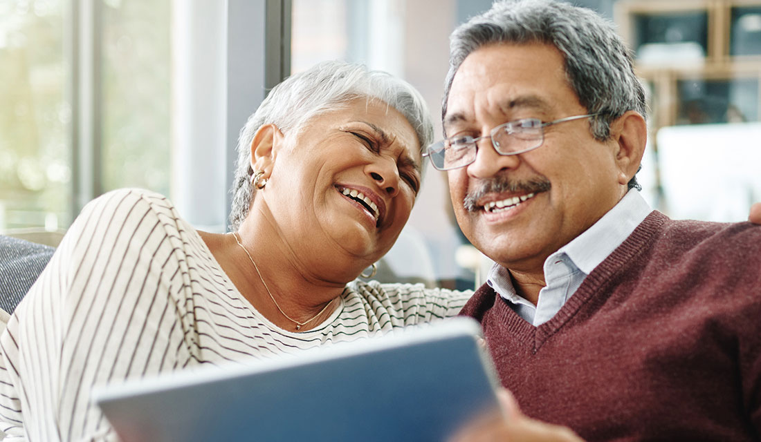Senior couple laughing together while looking at a tablet in their living room.
