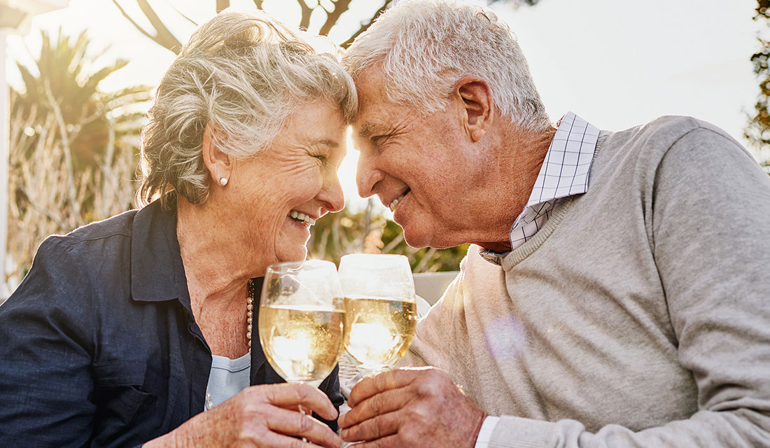 Elderly couple enjoying a sunny day with two glasses of champagne, smiling at each other.