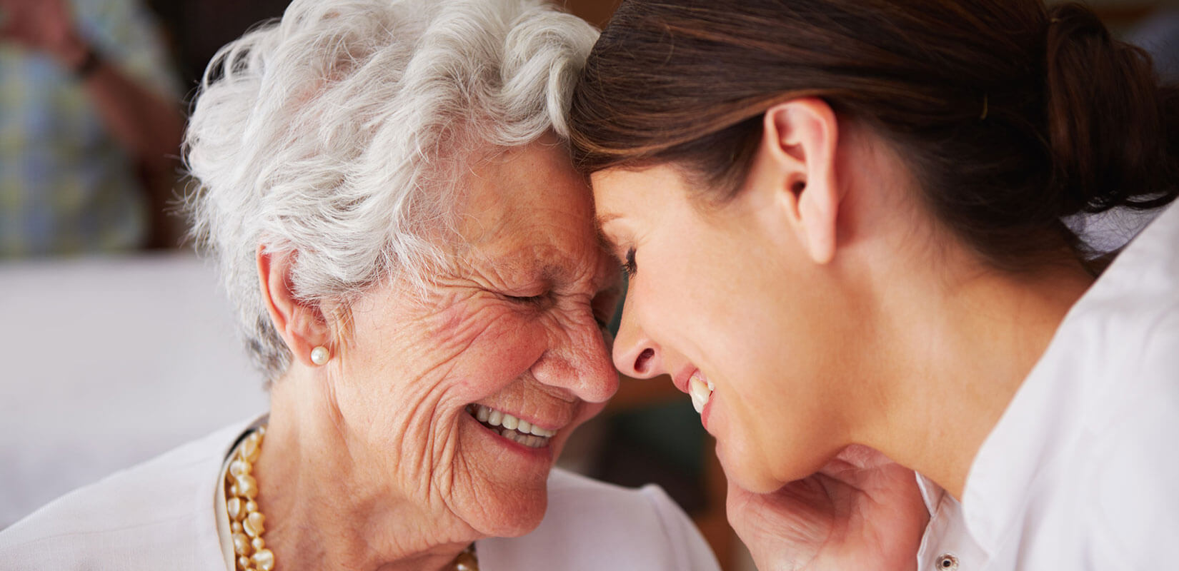 Elderly woman smiling with younger female caregiver. Foreheads touching affectionately.