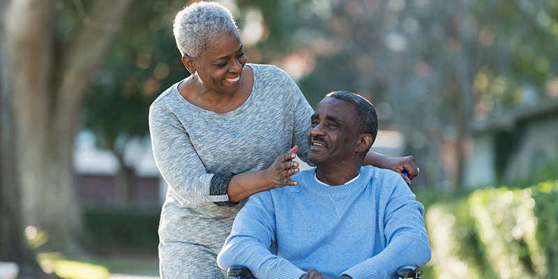 Senior woman smiling at man in wheelchair outdoors at senior living community.