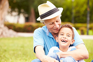 Elderly man in a hat and young boy laughing together while sitting on grass outside.