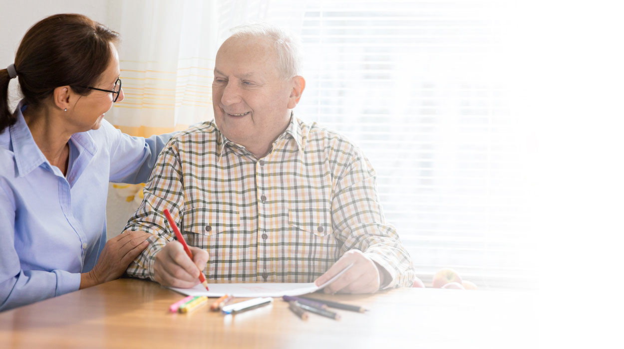 Elderly man in plaid shirt drawing with a red pencil as a female caregiver smiles beside him.