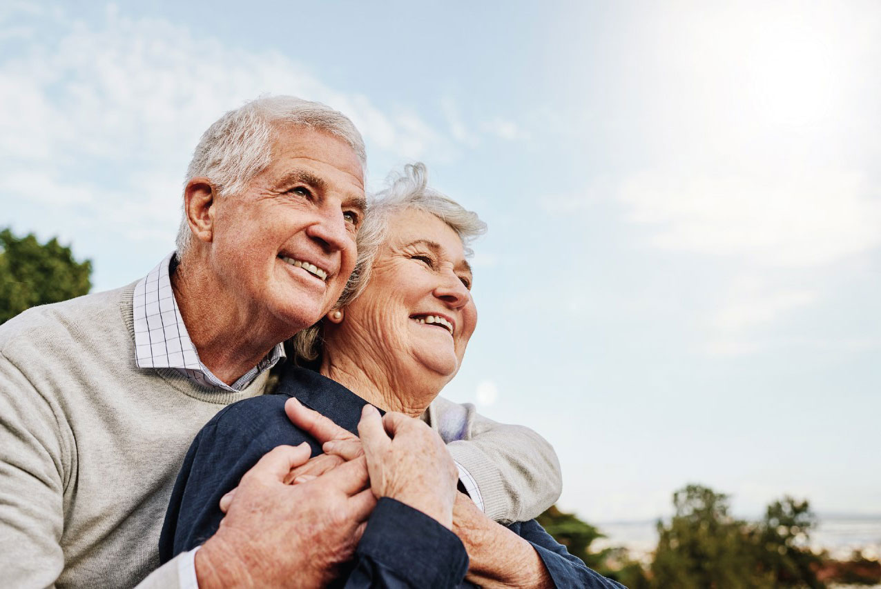 Smiling senior couple embracing outdoors with a bright sky in the background.