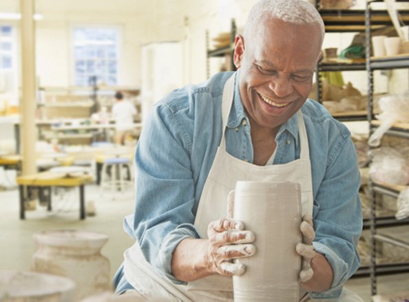 Smiling senior man in pottery class shaping clay with hands, shelves of ceramics in background.