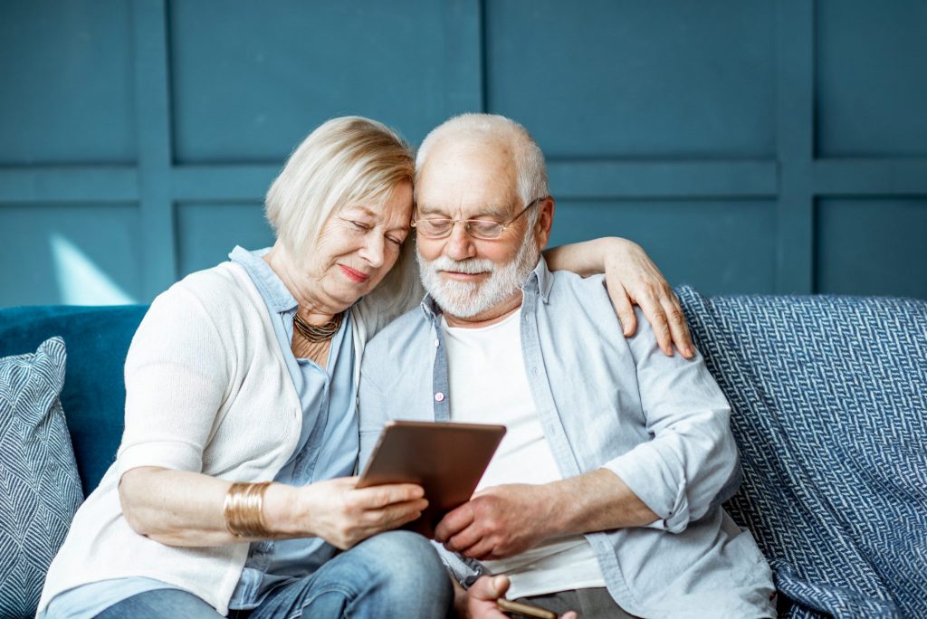 Senior couple seated on a blue sofa, smiling while looking at a tablet together.